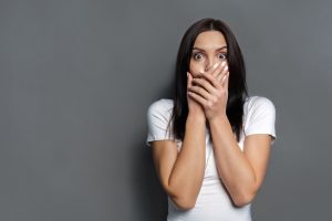 Keep silence. Scared woman covering mouth with hands while posing to camera on gray studio background. Shocked girl close lips with palms, speak no evil concept